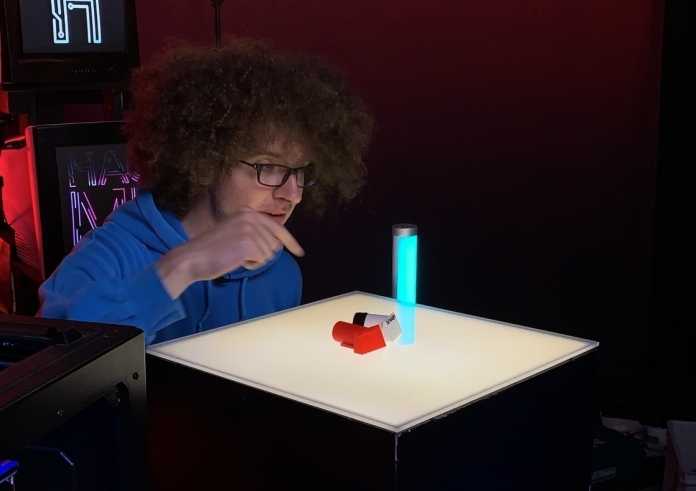 Ein junger Mann mit wilden Haaren schaut auf einen beleuchteten Tisch mit Teilen aus dem 3D-Drucker.