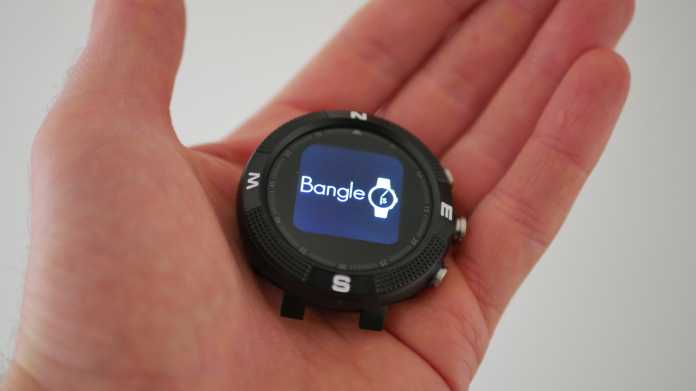 Eine Hand hält das runde Display einer schwarzen Smartwatch. Darauf steht: Bangle.js.