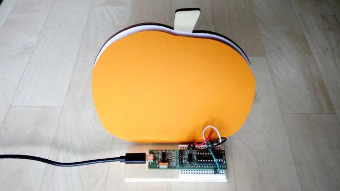 Eine orange Kürbisform, davor steht ein Breadboard mit grünem Mikrocontroller.