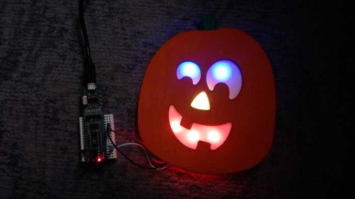 Ein dunkles Bild: Zu erkennen ist ein oranger Kürbis mit grün leuchtenden Augen und rot leuchtendem Mund, daneben ein Mikrocontroller auf einem Steckbrett.