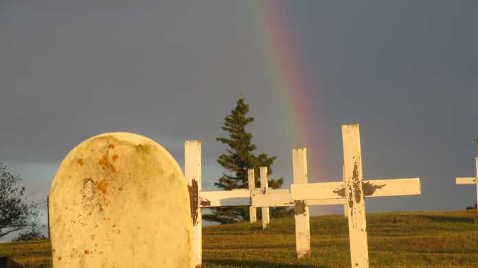 Weißer Grabstein, weiße Kreuze, dahinter ein Regenbogen