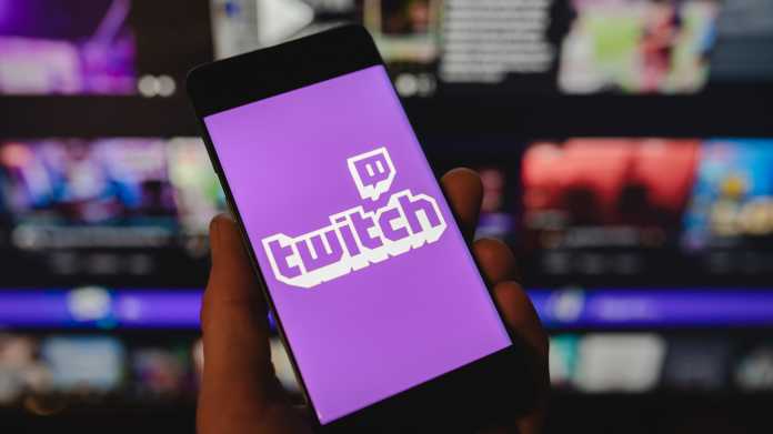 Smartphone mit dem Twitch-Logo auf dem Bildschirm vor einem verschwommenen Hintergrund mit mehreren Monitoren.