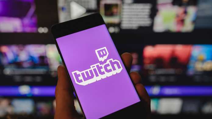 Smartphone mit dem Twitch-Logo auf dem Bildschirm vor einem verschwommenen Hintergrund mit mehreren Monitoren.