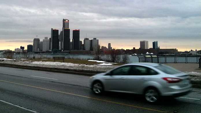 Blick auf die Skyline Detroits. Davor der Grenzfluss Detroit und ein vorbeifahrender Pkw