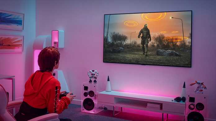 Eine sitzende Frau spielt ein Computerspielt, das auf einem an der Wand hängenden Fernseher läuft