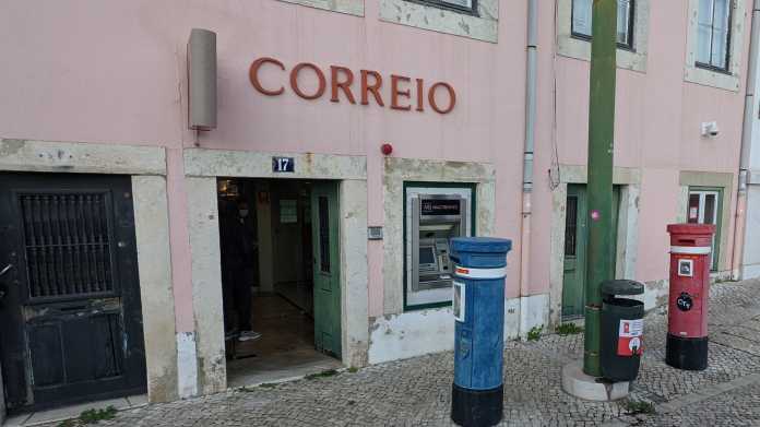 Rosa Gebäude mit Aufschrift &quot;Correio&quot;, über dem offenen Eingang eine vertikale Mobilfunkantenne, im Eingangsbereich ein maskierte Person, vor dem Haus zwei säulenförmige Briefkästen (blau bzw. rot) sowie ein Mistkübel, in der Gebäudefassade eingelassen ein Bankomat; eine maskierte Person steht im 