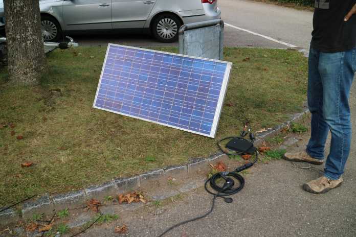 Solarmodul auf einer Grasfläche an ein Schild gelehnt, Wechselrichter und Kabel liegen im Vordergrund, eine Person steht daneben.