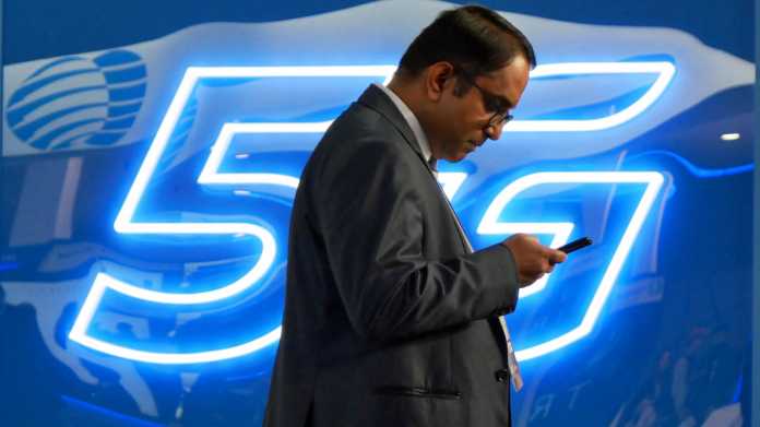 5G Symbolbild: "5G" in Leuchtshrift, davor ein Mann auf sein Handy blickender Mann