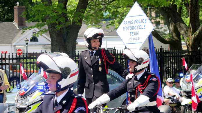 Mehrere Polizisten auf Motorrädern in Paradeuniform mit Helmen, eine Polizistin stehend auf ihrem Motorrad; dazu ein Schild "Paris Police Motorcycle Team" sowie kanadische und französische Wimpel auf den Motorrädern