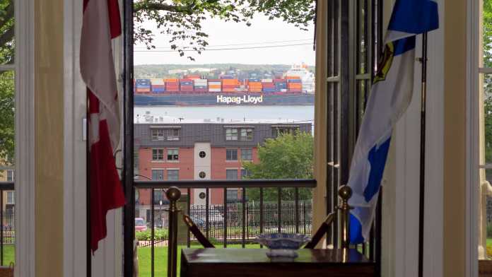 Durch ein Fenster sind Meerwasser und ein großes Containerschiff mit Aufschrift Hapag Lloyd zu sehen. Neben dem Fenster stehen links eine kanadische und rechts eine neuschottische Standarte, davor ein kleiner Tisch.