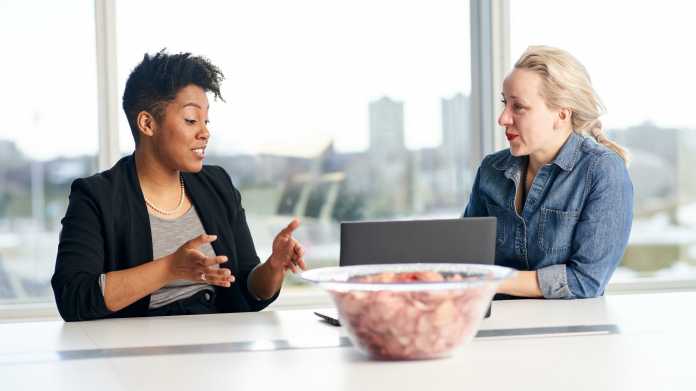 Zwei Frauen haben in einem Konferenzraum eine Besprechung, einen hat einen offenen Laptop; im Vorderung steht eine Schale Erdäpfelchips
