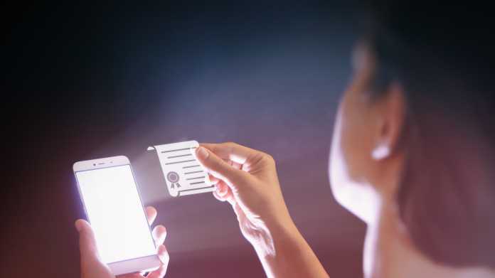 Frau hält einen Zettel vor ein Handy - der Zettel symbolisiert ein Zertifikat