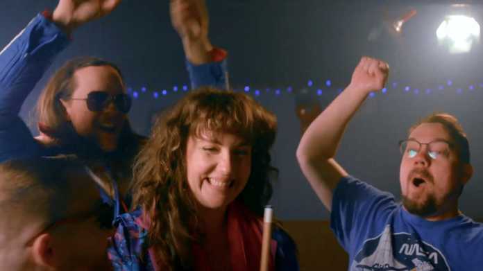 Szene aus Musikvideo: Lächelnde Frau mit Queue, dahinter feiernde Männer