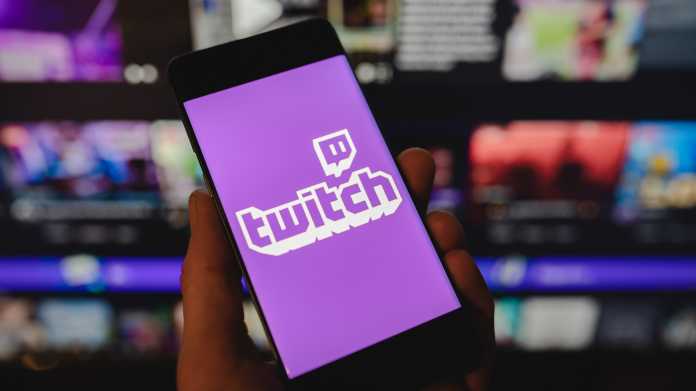 Smartphone mit dem Twitch-Logo auf dem Bildschirm vor einem verschwommenen Hintergrund mit mehreren Monitoren.