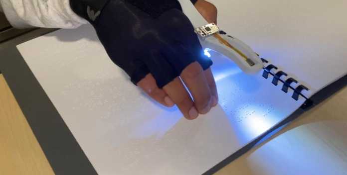 Eine Person liest Brailledruck; zwischen Daumen und Zeigefinger hält sie ein Gerät, das ein helles Licht auf das Papier strahlt