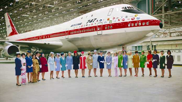 Historische Aufnahme: Eine weiße Boeing 747 mit einem roten Streifen; davor stehen 26 schlanke Frauen in unteschiedlichen Air-Hostess-Uniformen.