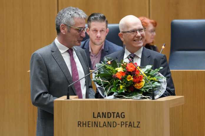 Hendrik Hering und Dieter Kugelmann lachend im Landtag Rheinland-Pfalz am Rednerpult, auf dem ein Strauß Blumen liegt