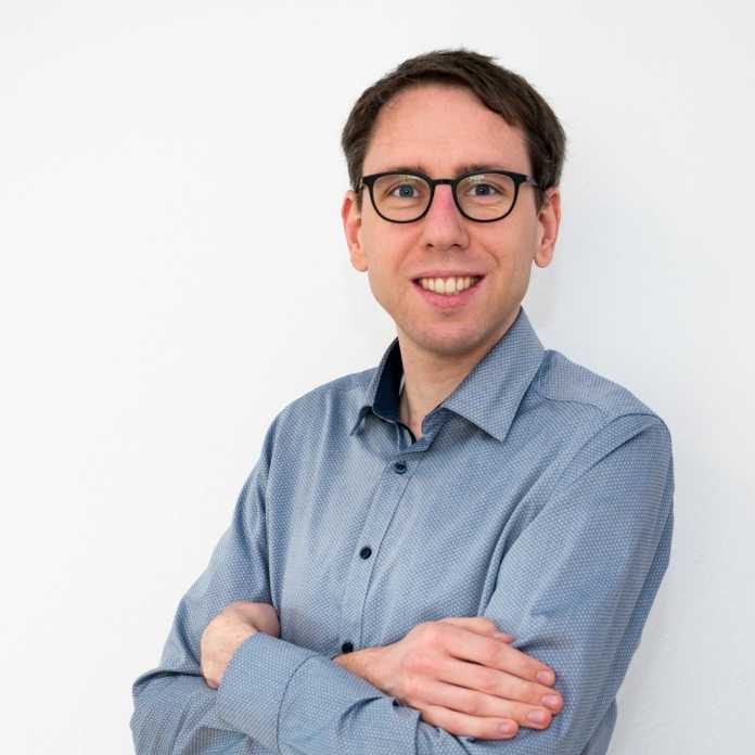 Porträt eines weißen Mannes mit Brille, verschränkten Armen, graublauem Hemd