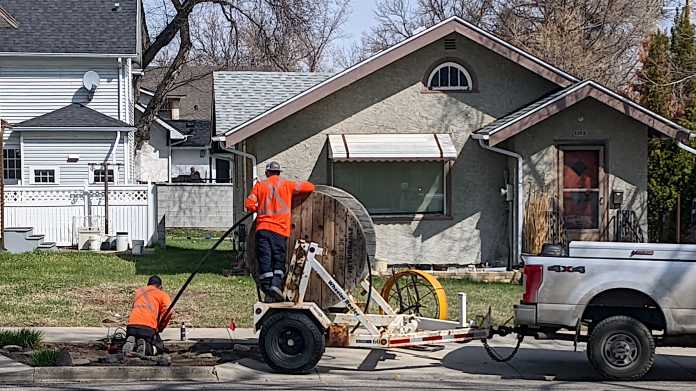 Zwei Männer in orange Overalls führen Glasfaser von einer hölzernen Kabelrolle in einen Schacht; im Hintergrund Einfamilienhäuser und Bäume