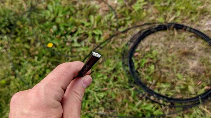 Eine Hand hält das abgeschnittene Ende eines Glasfaserkabels; am unscharf zu sehenden Wiesenboden liegt eine Rolle des Kabels.