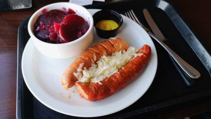 Auf einem Tablett steht ein Teller mit zwei Würste, Sauerkraut und einer Schale Rote-Rüben-Salat, dazu Senf sowie Messer und Gabel