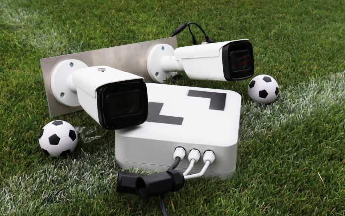 Auf dem Rasen liegt ein Metallblech mit zwei weißen Kameras und ein kleines Computergehäuse, dazu zwei kleine Bälle