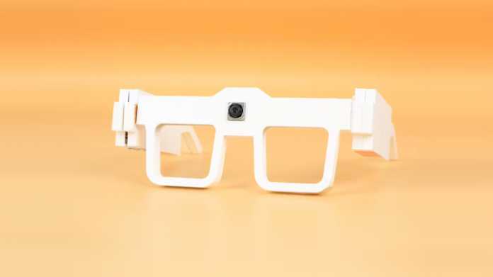 Eine weiße, 3D-gedruckte Brille vor orangem Hintergrund. In der Mitte zwischen den Linsen ist eine Kamera zu sehen.