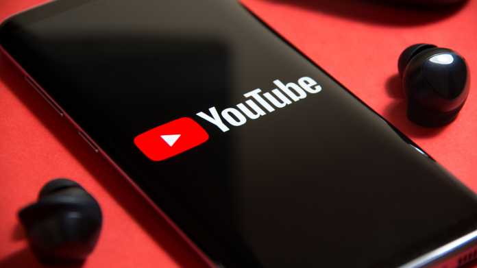 Handy, auf dem Youtube-Logo und -Schriftzug zu sehen sind; links und rechts davon liegt jeweils ein schwarzer, kabelloser In-Ohr-Kopfhörer