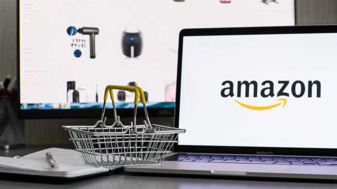 Miniatureinkaufskorb links neben einem Laptop mit Amazon-Logo. Im Hintergrund die Amazon Buy Boxes, in denen Produkte auf der Website des Online-Gigangten Amazon platziert sind.