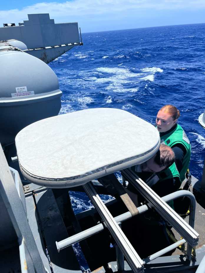 Flache, weiß, quadratische Antenne, die von 2 Personen auf einem Metallgestände angebracht wird; im Hintergrund Meerwasser
