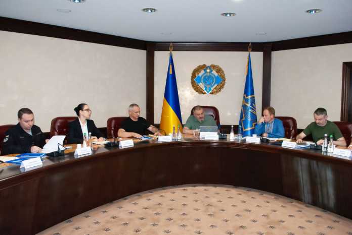 5 Männer und 1 Frau sitzen an einem großen, ovalen Tisch; dahinter 2 Flaggen und ein Wappen an der Wand