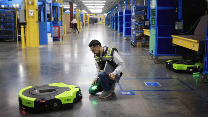 A white man in a high-visibility vest kneels on the floor of a warehouse next to a roundish, flat robot that resembles an oversized vacuum cleaner robot.