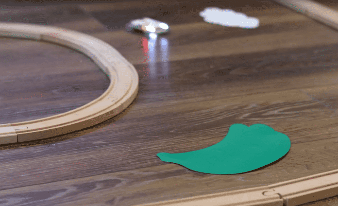 Man sieht die Kurve einer kleinen Rennstrecke. Die Banden sind aus Holzeisenbahnschienen gebaut. Von hinten im Bild fährt ein kleines Auto Richtung Kamera. Weiter vorn auf der Strecke liegt ein grünes, flach auf dem Boden liegendes Hindernis.