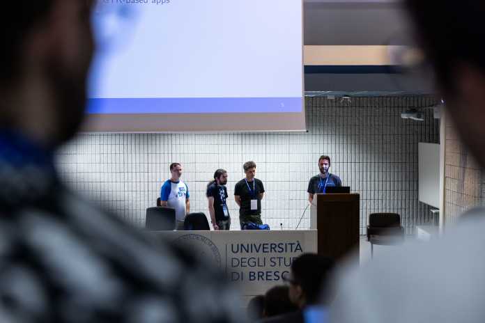 Three rather younger men are standing on the podium in a large lecture hall, the fourth, Sebastian Wick, at the lectern.
