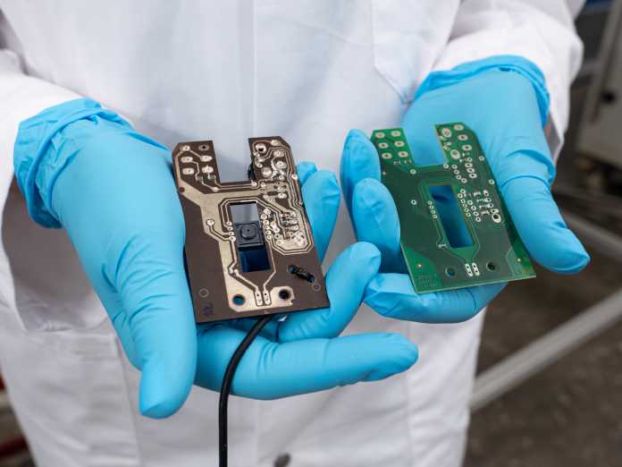 Blue-gloved hands hold a degradable and a conventional circuit board in the camera