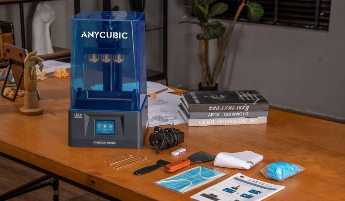 Auf einem braunen Tisch stehen ein 3D-Drucker mit blauer Verkleidung und Zubehörteile wie Netzteil und Spatel.