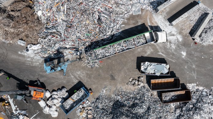Luftaufnahme eines Aluminium-Recyclingbetriebs mit Metallschrotthalden, Schüttmulden, Lastwagen und einem Ladebagger.
