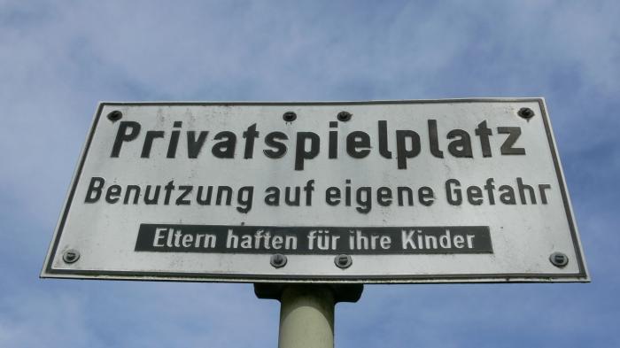 Ein Schild vor blauem Himmel, die Aufschrift lautet: Privatspielplatz - Benutzung auf eigene Gefahr - Eltern haften für ihre Kinder