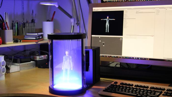 Vor einem Bildschrim steht ein schwarzes Gehäuse, in dessen Licht steht eine "holografische" Frauenfigur