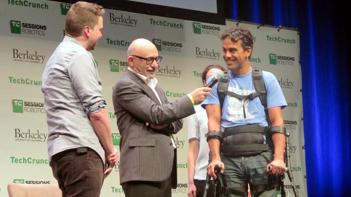 3 Männer auf einer Bühne, davon einer mit Krücken und Exoskelett