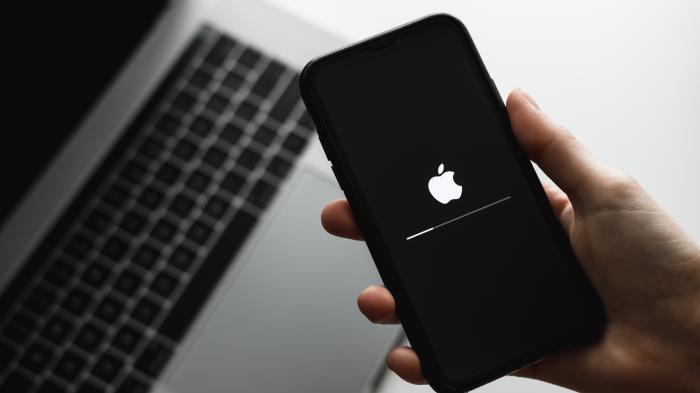 Eine Hand hälte ein iPhone, auf dem gerade ein Update läuft, darunter steht ein geöffnetes MacBook