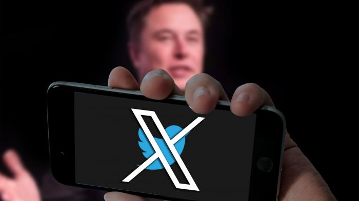 Unscharfer Elon Musk hinter Smartphone mit Logo von X vor Twitter-Vogel