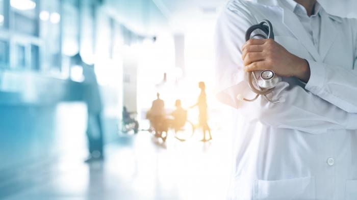 A doctor in a hospital corridor. His arms are crossed. Behind him you can see blobs that could be people.