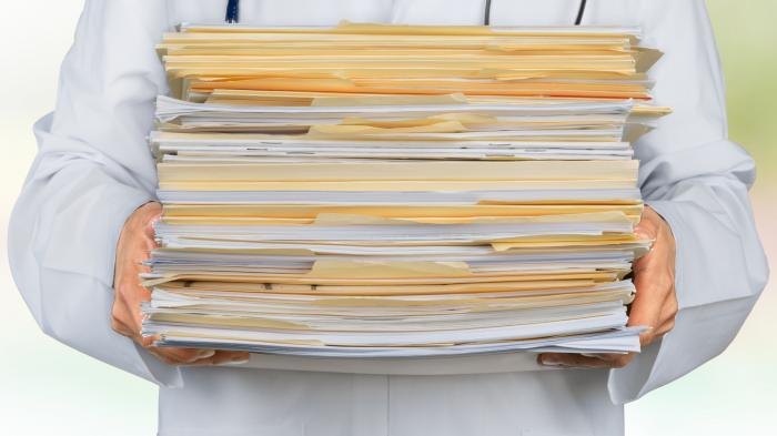 A doctor with a stethoscope around his neck holds a pile of paper. All that can be seen of the doctor is his upper body in a white coat and two hands carrying the pile of paper.