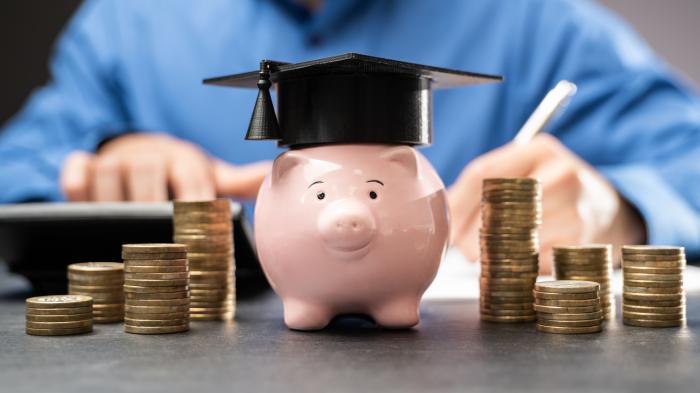 A small glass with coins, on which lies a cardboard replica of a graduate's hat, on a pile of school books. A blackboard in the background.