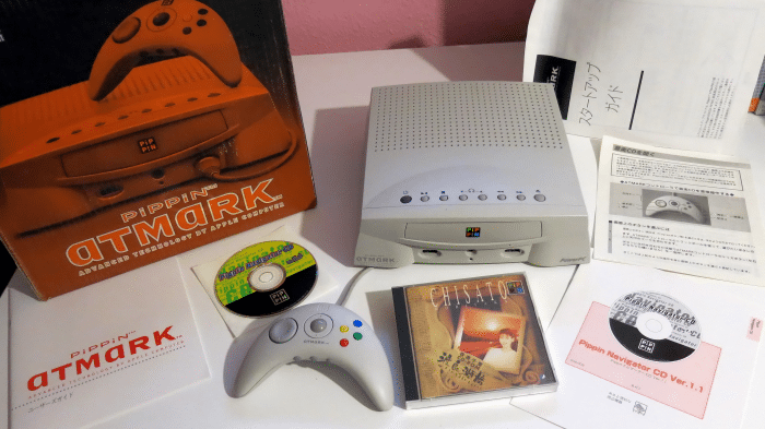 Die Spielekonsole „Pippin Atmark“ von Bandai im vollen Lieferumfang. Sie ist grau und hat Tasten für CD und Lautstärke an der Oberseite, an der Front CD-Schublade und Controllerports.