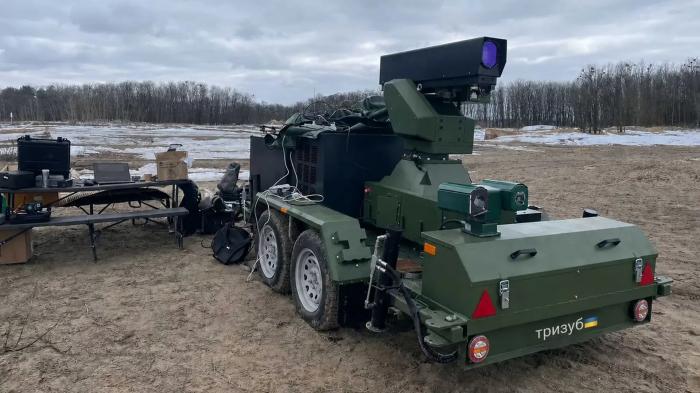 Ein militärisches Lasersystem auf einem Anhänger steht im Freien neben einem Tisch mit Laptops.