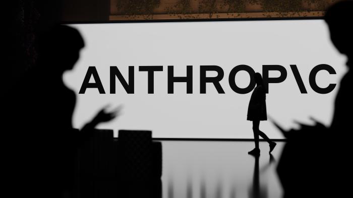 Anthropic-Schriftzug auf einem riesigen Bildschirm, vor dem eine Person vorbeigeht; im Vordergrund sitzen links und rechts je eine Person
