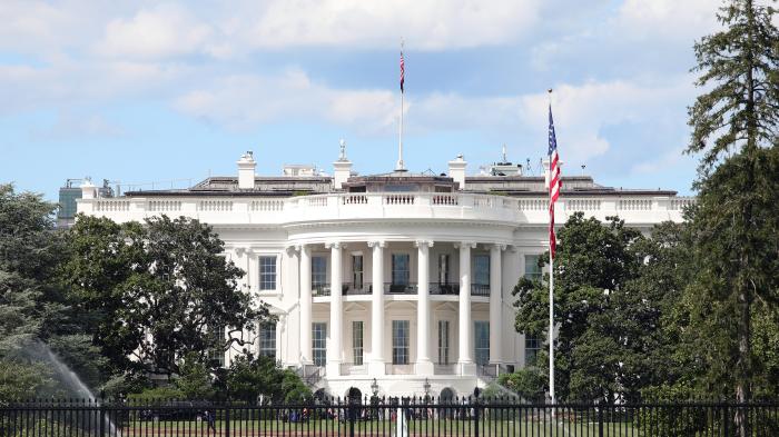 Weißes Haus in Washington D.C., Frontansicht mit Säulen, US-Flagge, Garten und Zaun im Vordergrund bei Tageslicht
