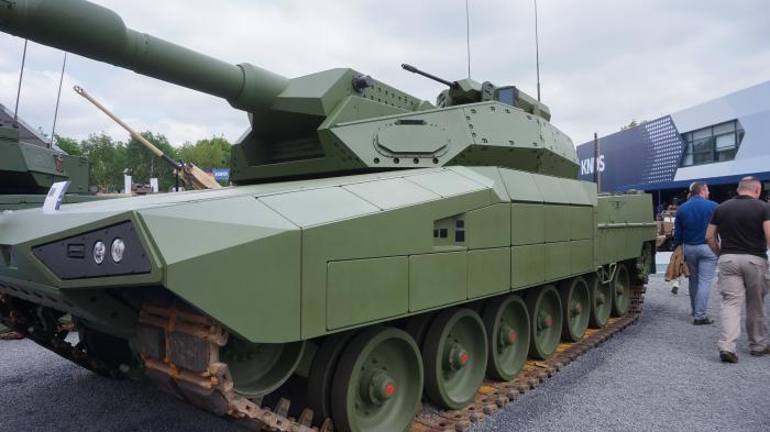 Ein grüner Panzer mit Ketten und einer langen Kanone steht auf Schotter.